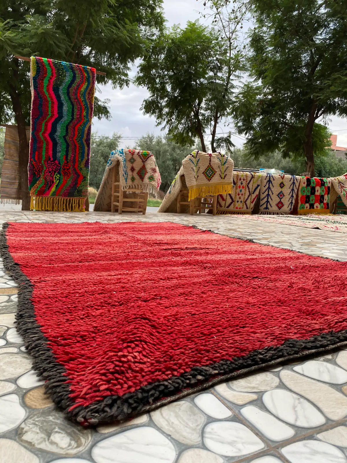 Whole red shaggy rug with a dark brown edge laid on a patio, with patterned rugs draped on chairs behind