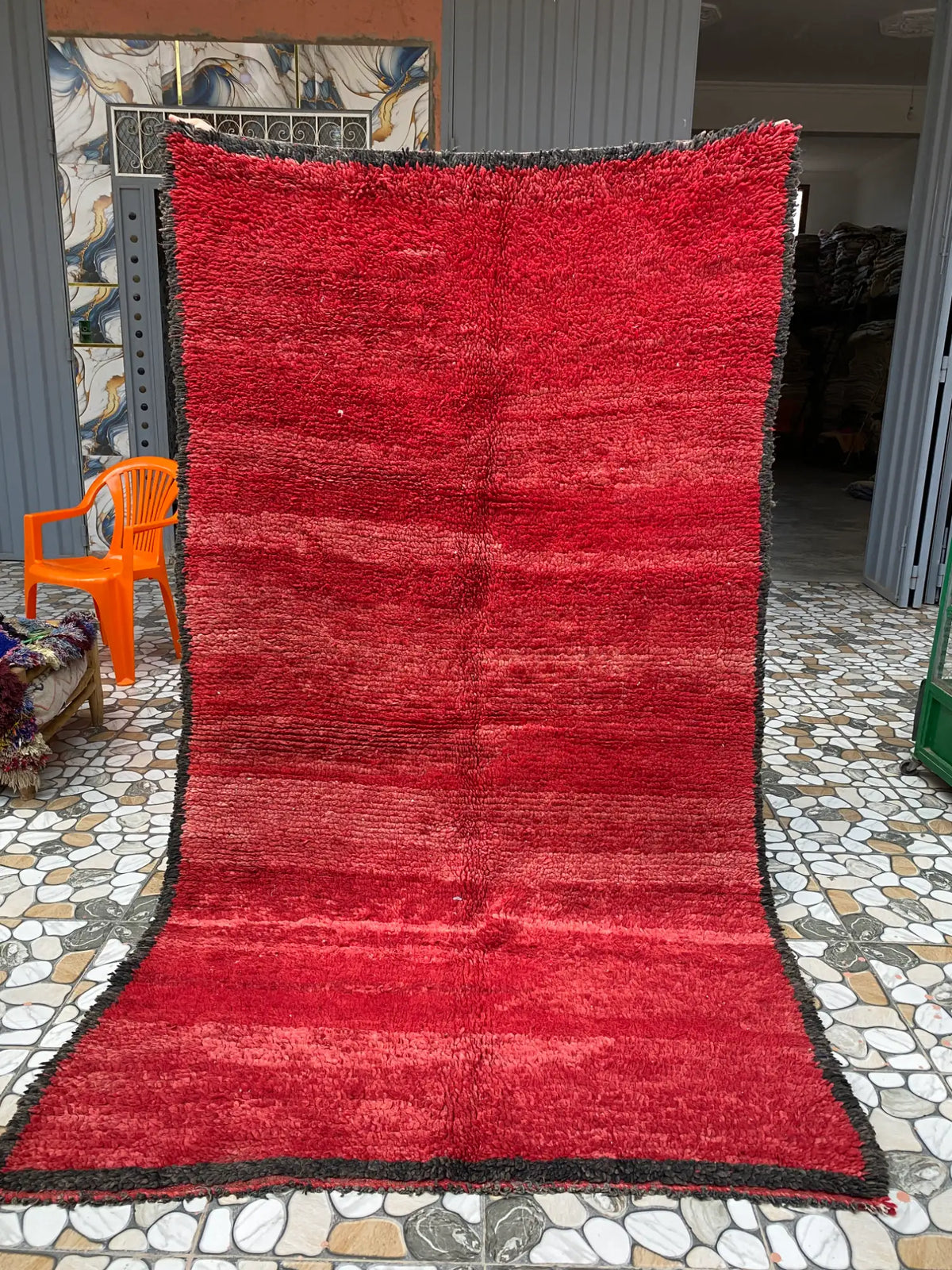 Red shaggy rug with dark gray edging held upright outdoors on a mosaic tile floor in front of a doorway