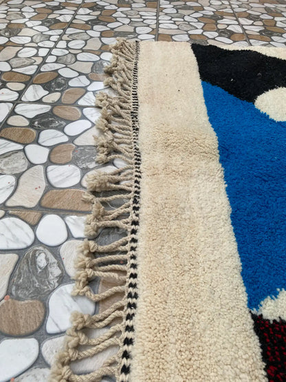 Close-up of the rug's cream pile, black and white braided edge and knotted fringe beside a blue shape