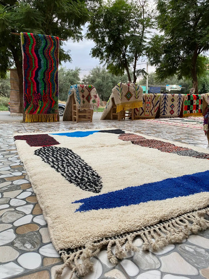Cream shaggy rug with blue, black and rust abstract shapes on a stone-tiled patio, other rugs displayed behind