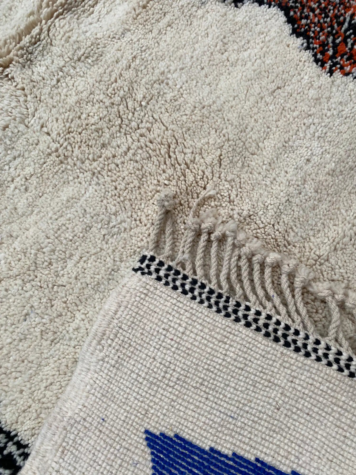 Close-up of the soft cream wool pile and knotted fringe, with the rug's back edge folded over to show black stitching