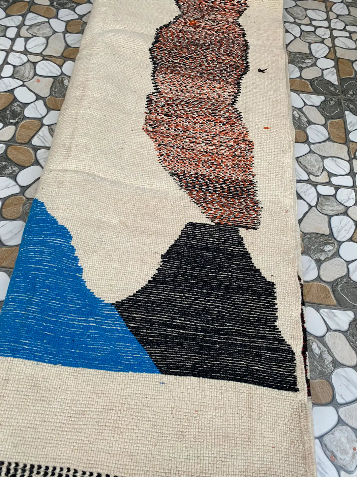 Back of the rug showing the flat woven cream base with blue, black and rust-brown shapes on a pebble-tiled floor