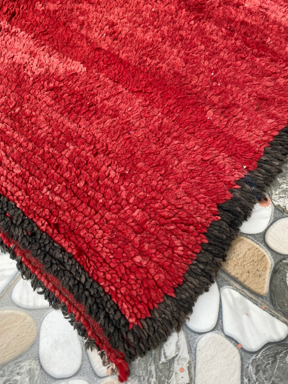 Corner of a red shaggy rug with a dark brown woolly border and red fringe, lying on pebble-pattern tiles